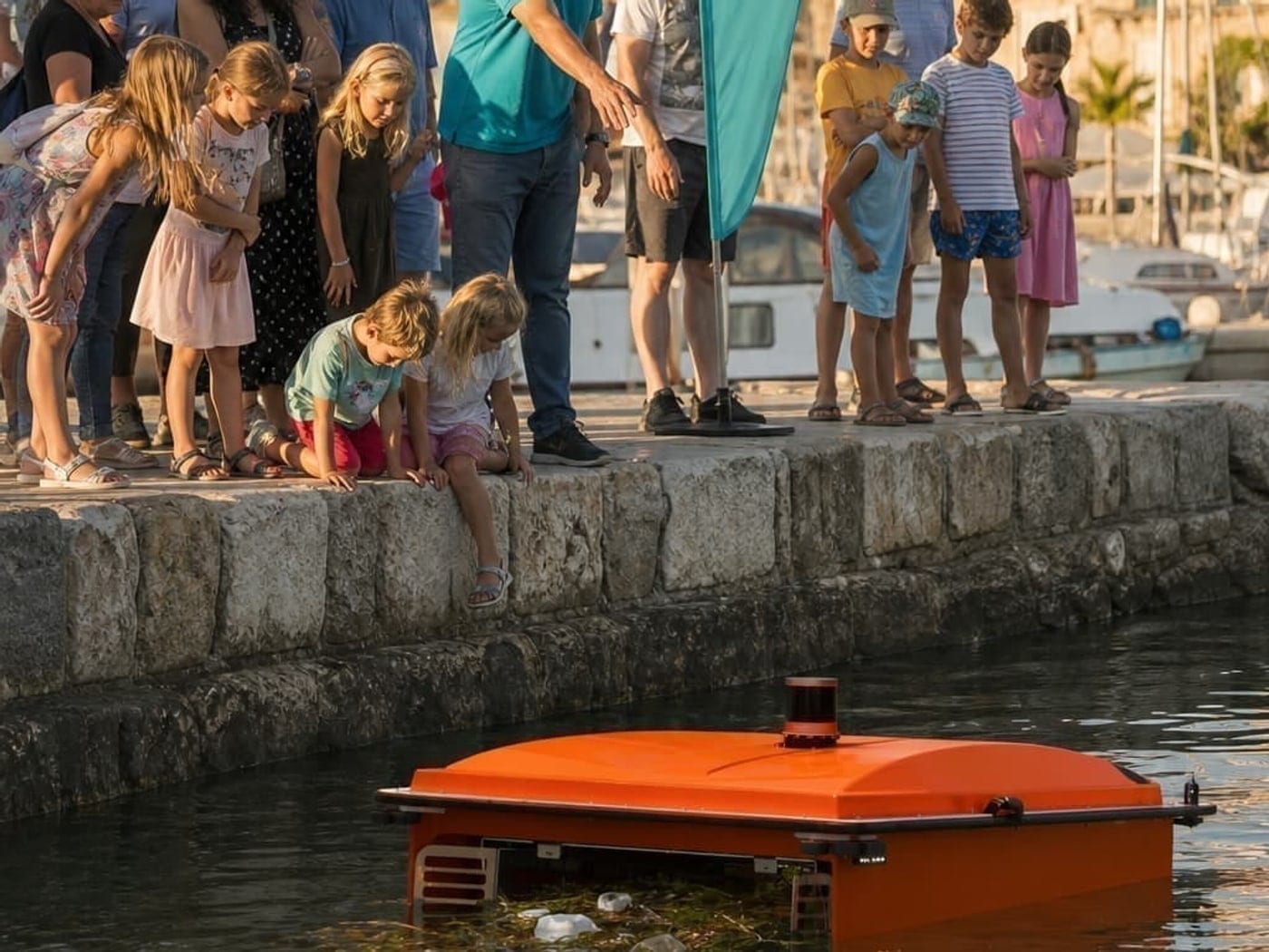 A crowd of families on a harbor wall watching a WasteShark drone collect litter below them